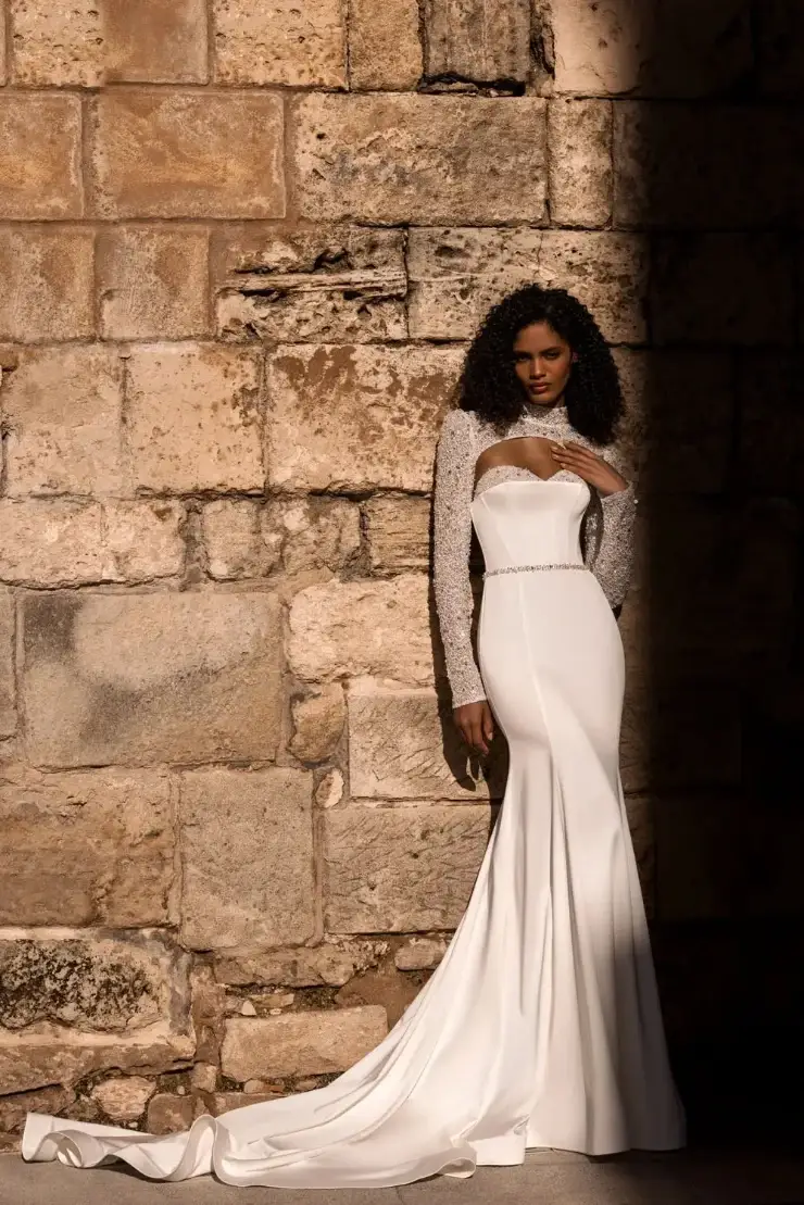 Elegant woman in a fitted, strapless white bridal gown stands confidently in a sunlit archway. The setting is warm and inviting, conveying grace.